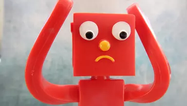 A personified red toy, rubber-like, with a square head, arms raised, and a distressed look like being confused or distressed
